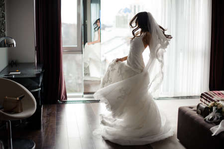 Handsome elegant brunette bride in white lacy wedding dress and veil dancing near the windowの写真素材