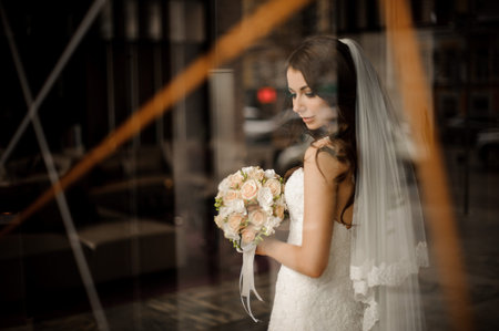 beautiful charming bride in white wedding dress with tender bouquet of roses mysteriously smiles, looking out of the windowの写真素材