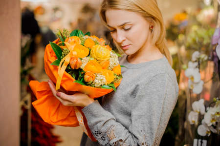 blonde girl in gray sweater holds a bouquet of orange roses and tulips in orange packingの写真素材