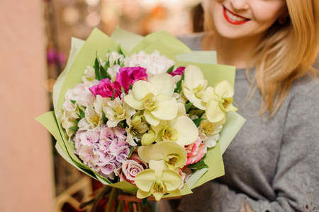 blonde female in gray sweater smiles and holds a bouquet with beige orchids, pink roses and purple hydrangea in packingの写真素材