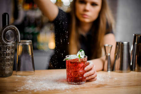 pretty professional barista finishes preparation of pink alcoholic cocktail with mint leaves by adding a bitter of powdered sugarの写真素材