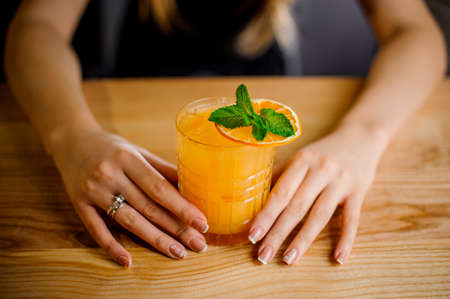 barmaid holds a crystal glass with a cocktail of orange color decorated with a sprig of mint and a slice of dried orangeの写真素材