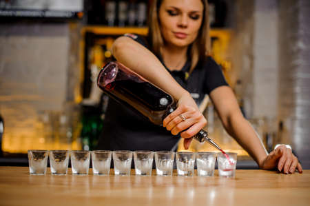 beautiful barmaid pours brightly crimson alcohol from a bottle into crystal stacks that are lined upの写真素材