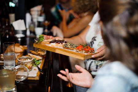 Waitress hands holding a big wooden plate with different kinds of sandwiches near the tableの写真素材