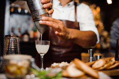 Barman dressed in a brown leather apron pouring alcoholic cocktail into the glass standing behind the bar counterの写真素材