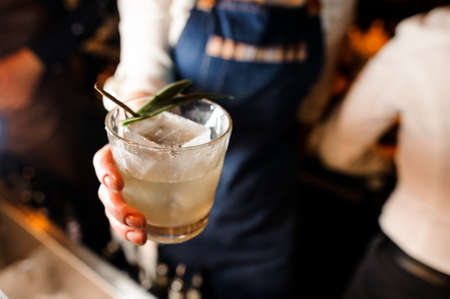 Waitress dressed in a white shirt and blue apron holding a glass filled with cold alcoholic drink with ice cubesの写真素材