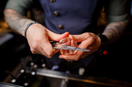 barman with tattoos on his hands cuts the ice with a special knifeの写真素材