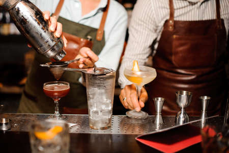 two barmen in aprons at the bar counter prepare alcoholic cocktail with ice , using shaker and strainerの写真素材