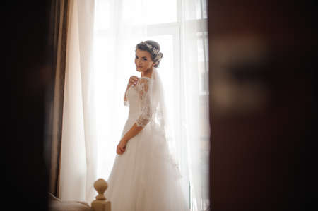beautiful brunette bride in a white lace dress stands on a window background in a bedroomの写真素材