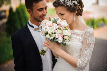 beautiful bride with a pink manicure, a lace dress and a gentle styling enjoys the scent of a wedding bouquet, the groom stands beside smilingの写真素材