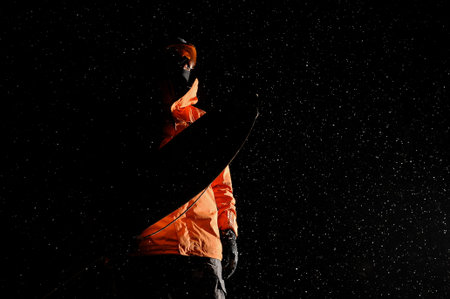 Bottom view of darkened figure of a male snowboarder standing with a snowboard at the dark snowy...