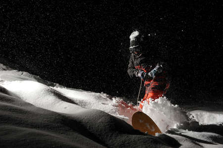 Snowboarder dressed in the orange sportswear riding with the snowboard at the dark snowy nightの写真素材
