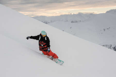 Freeride male snowboarder in sportswear sliding down the snowy slope on the background of sunset skyの写真素材