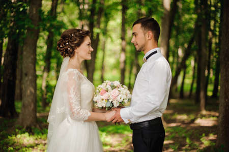Young and beautiful bride and bridegroom look and holding one another hands on the background of green forestの写真素材
