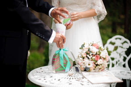 Bridegroom with bride pouring multi-colored sand under the white table against the blurred green backgroundの写真素材