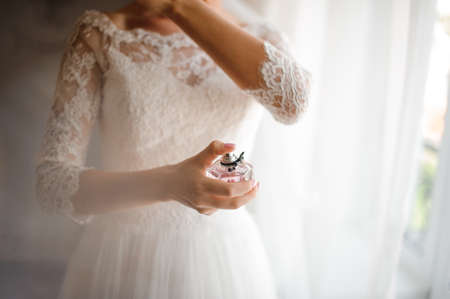 Bride in a beautiful white wedding dress holding a perfume and standing against the background of the windowの写真素材