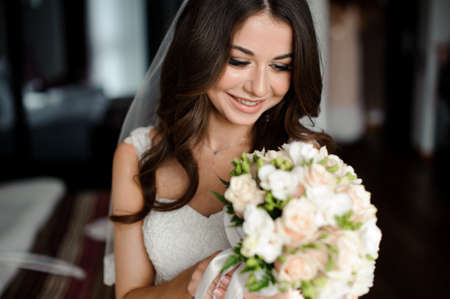 Bride morning preparation. Beautiful and smiling bride in a white veil holding a lovely wedding bouquet of flowersの写真素材