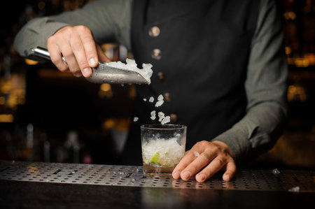 Bartender adding ice cubes into the cocktail glass with pieces of lime making Caipirinha cocktailの写真素材