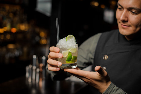 Smiling barman holding a glass filled with fresh and cool Caipirinha cocktail with a straw
