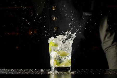 Glass filled with splashing fresh Caipirinha cocktail on the bar counter against the dark background...
