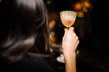 Back view of brunette woman holding an elegant glass filled with alcoholic cocktail on the dark backgroundの写真素材