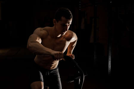 Sexy and young muscular shirtless man practicing crossfit in a dark gym using special trainersの写真素材