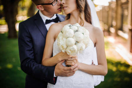 stylish groom with glasses is going to kiss the charming bride in a white dress, with a veil and a bouquet of peoniesの写真素材