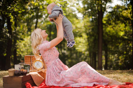 Blonde mother dressed in a dress playing with a baby on a picnic in the forestの写真素材