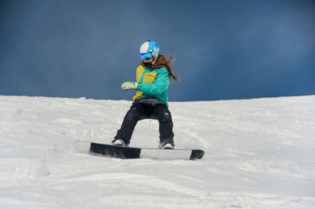 Female snowboarder dressed in sportswear riding down the mountain slope on the bright sunny dayの写真素材