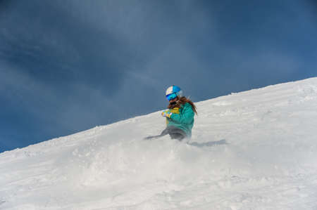 Female snowboarder dressed in sportswear and helmet riding down the mountain slope on the bright sunny dayの写真素材