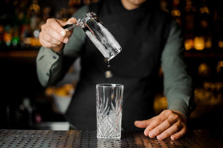 Bartender putting a big ice cube into an empty glass on the steel gray bar counter close-upの写真素材