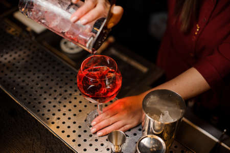 Professional bartender girl pouring a light red cocktail into the glass with ice at the steel bar counterの写真素材