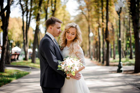 Portrait of a young bridegroom embracing a lovely blonde bride holding a beautiful bouquet of flowers in the parkの写真素材