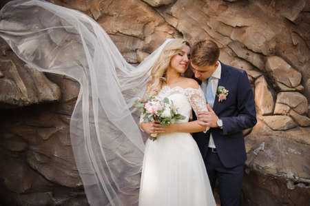 Portrait of a young bridegroom embracing a lovely blonde bride holding a bouquet of flowers on the background of wild stoneの写真素材
