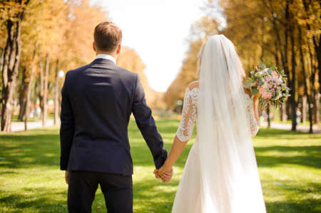 Back view of bride and bridegroom holding hands on the park lawn against the background of yellow treesの写真素材