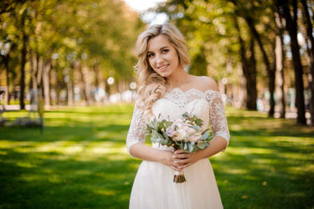 Lovely blonde bride dressed in a beautiful wedding dress holding a tender bouquet of flowers standing on the green lawnの写真素材