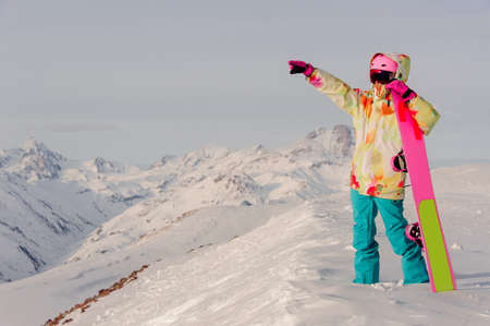 Female snowboarder wearing colorful jacket and blue pants standing with snowboard in one hand and enjoying mountain landscape in Gudauri, Georgiaの写真素材