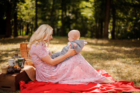 Blonde mother dressed in a dress playing with baby on a picnic in the forestの写真素材