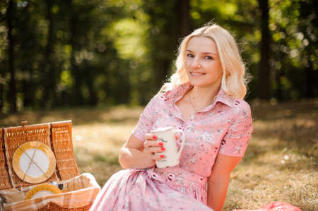 Lovely and young blonde woman sitting and holding a cup on picnic in the forestの写真素材