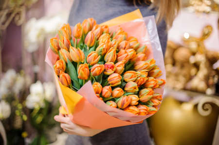Woman holding a large bright bouquet of orange tulips wrapped in a paper on the background of flower shopの写真素材