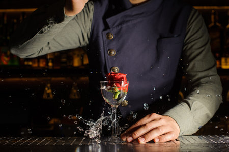 Elegant cocktail glass with splashing drink and rose in it on the bar counter against the barmanの写真素材
