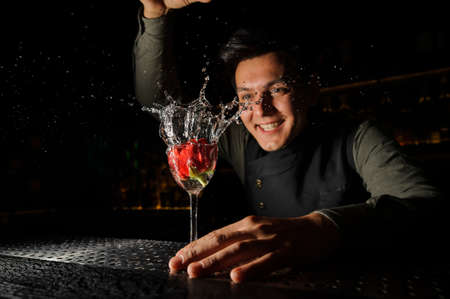 Smiling barman holding an elegant cocktail glass with splashing alcoholic drink and rose in it in on the bar counterの写真素材
