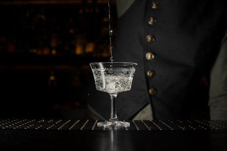 Barman pouring a transparent alcoholic drink into the cocktail glass on the dark bar counter