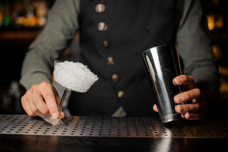 Barman hands holding a shaker and a cocktail glass filled with ice on the bar counter