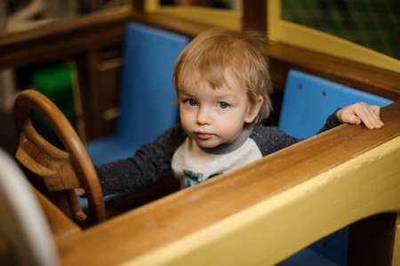 Cute little boy playing in the big toy wooden car with blue seats at the playing roomの写真素材