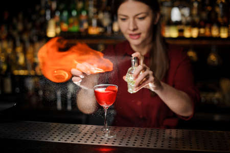 Young female bartender sprinkling a cocktail glass with tasty Aperol syringe cocktail with a peated whisky and making a smoky noteの写真素材