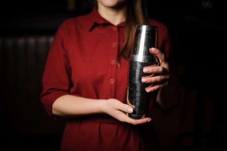 Professional bartender girl holding in hands a steel shaker with a fresh cocktail dressed in the red shirtの写真素材