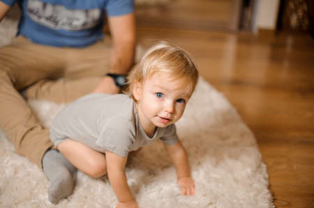 Cute blonde and blue-eyed baby dressed in a grey romper sitting on the carpet with fatherの写真素材