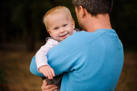 Father holding in hands and hugging a pretty and smiling blonde baby girl in parkの写真素材