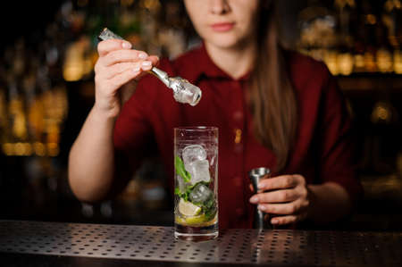 cute barmaid in a red dress prepares a mojito in a crystal glass, adding iceの写真素材